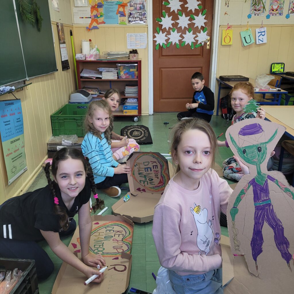 widzimy grupę uczniów trzymających swoje kolorowe prace wykonane z pudełek po pizzie