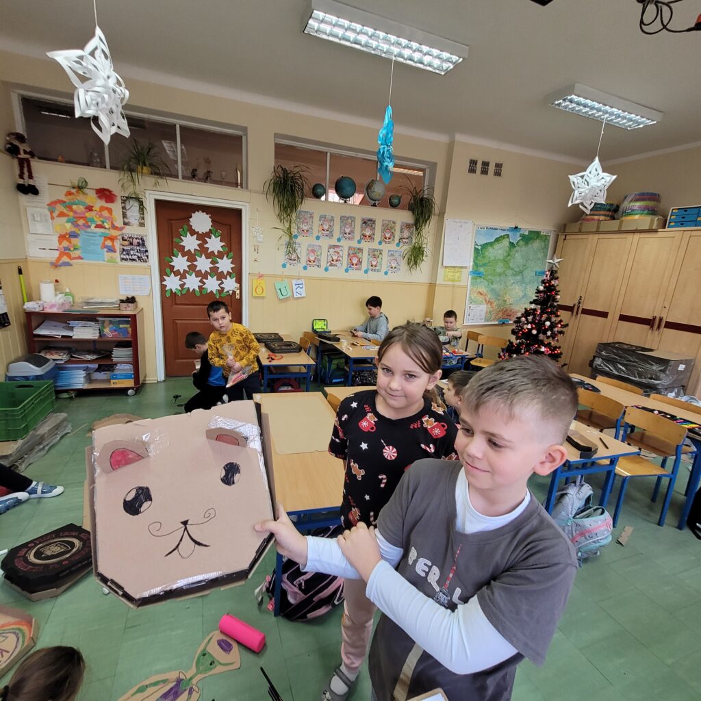 widzimy grupę uczniów trzymających swoje kolorowe prace wykonane z pudełek po pizzie