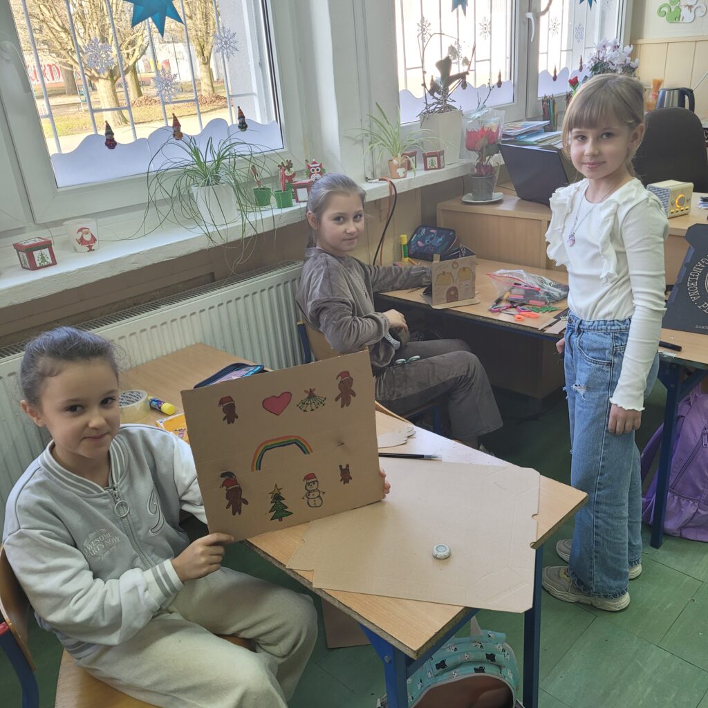 dziewczynka trzyma pracę. Malunek na wycinku pudełka. Obok stoi jedna koleżanka i druga siedzi.