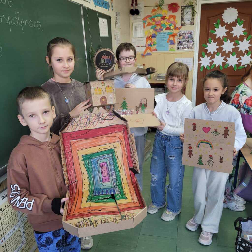 widzimy grupę uczniów trzymających swoje kolorowe prace wykonane z pudełek po pizzie