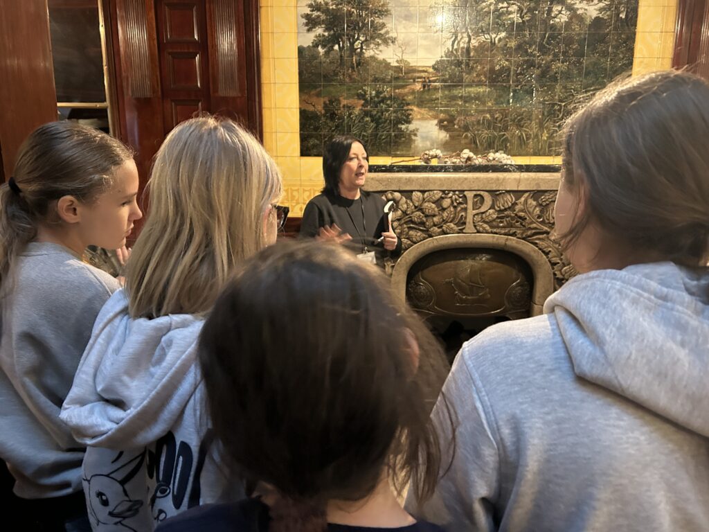 kilkoro dzieci stoi tyłem do aparatu , przodem do pani przewodnik, która dzieciom opowiada historię Łodzi