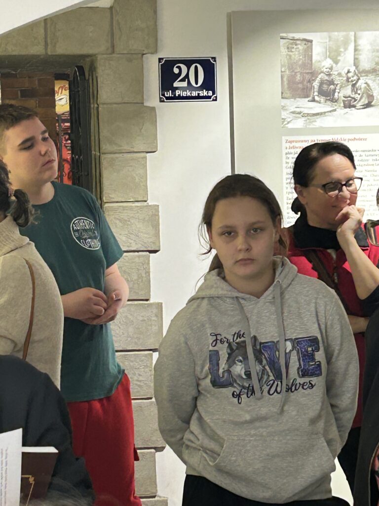 dziewczynka i chłopiec oraz nauczycielka przyglądają się wystawie, w tle tabliczka na ścianie ul. Piekarska 20