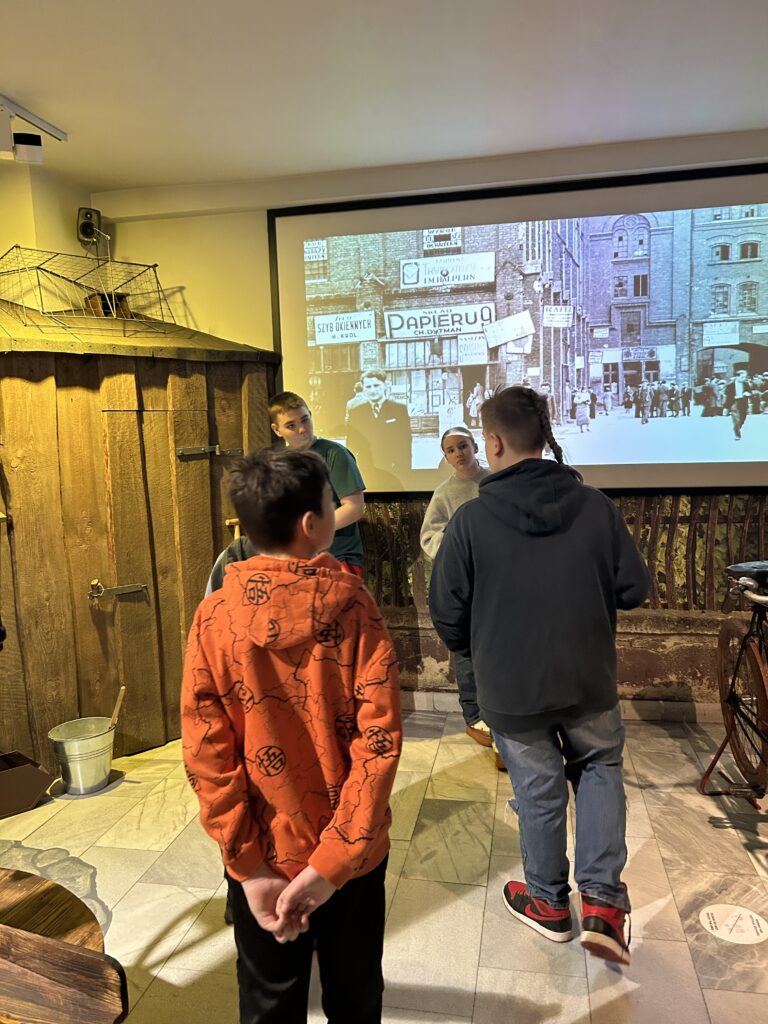 czwórka dzieci ogląda częśc wystawy poświęconej podwórkom  łódzkim. Jest drewutnia, wiaderko, widać część roweru. W tle film dokumentalny.