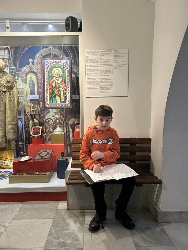 chłopiec siedzi na ławce i rozwiązuje zadania. Obok niego część wystawy poświęconej prawosławiu