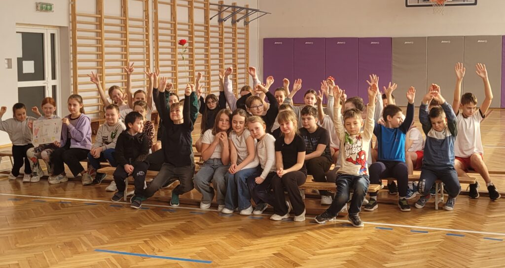 zdjęcie grupowe uczniów, siedzą na ławkach, trzymają ręce ku górze 