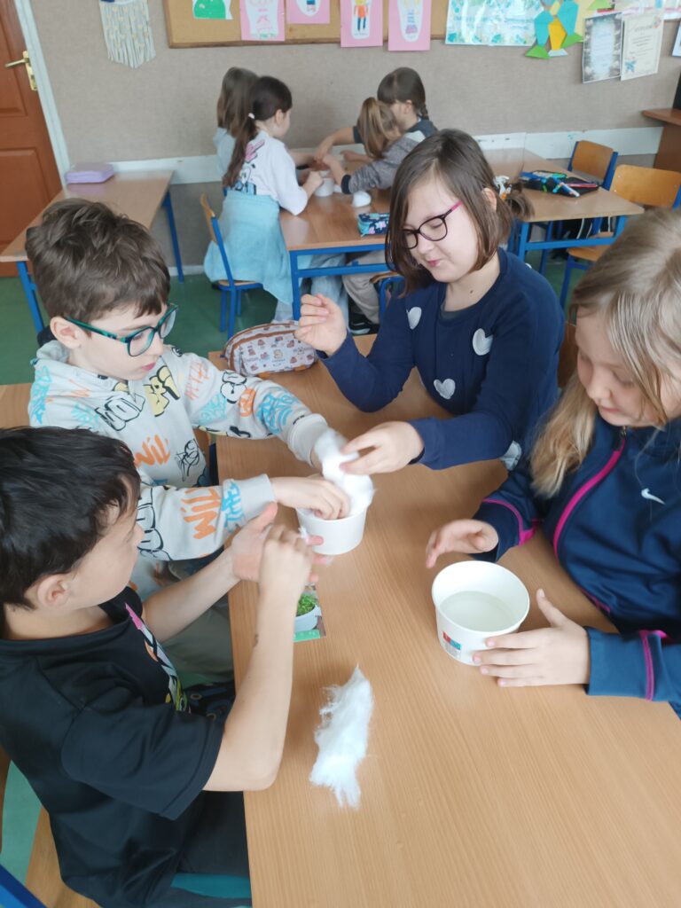 uczniowie siedzą grupą przy ławce, wsadzają ziarenka rzeżuchy do kubeczków  