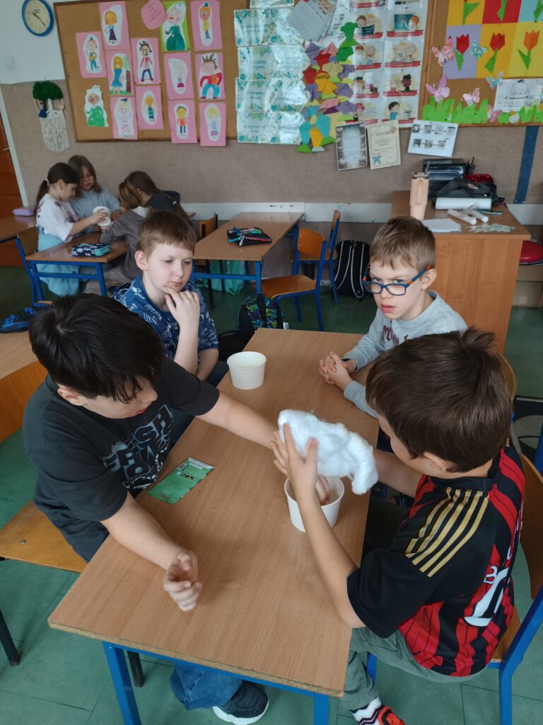 uczniowie siedzą grupą przy ławce, wsadzają ziarenka rzeżuchy do kubeczków  