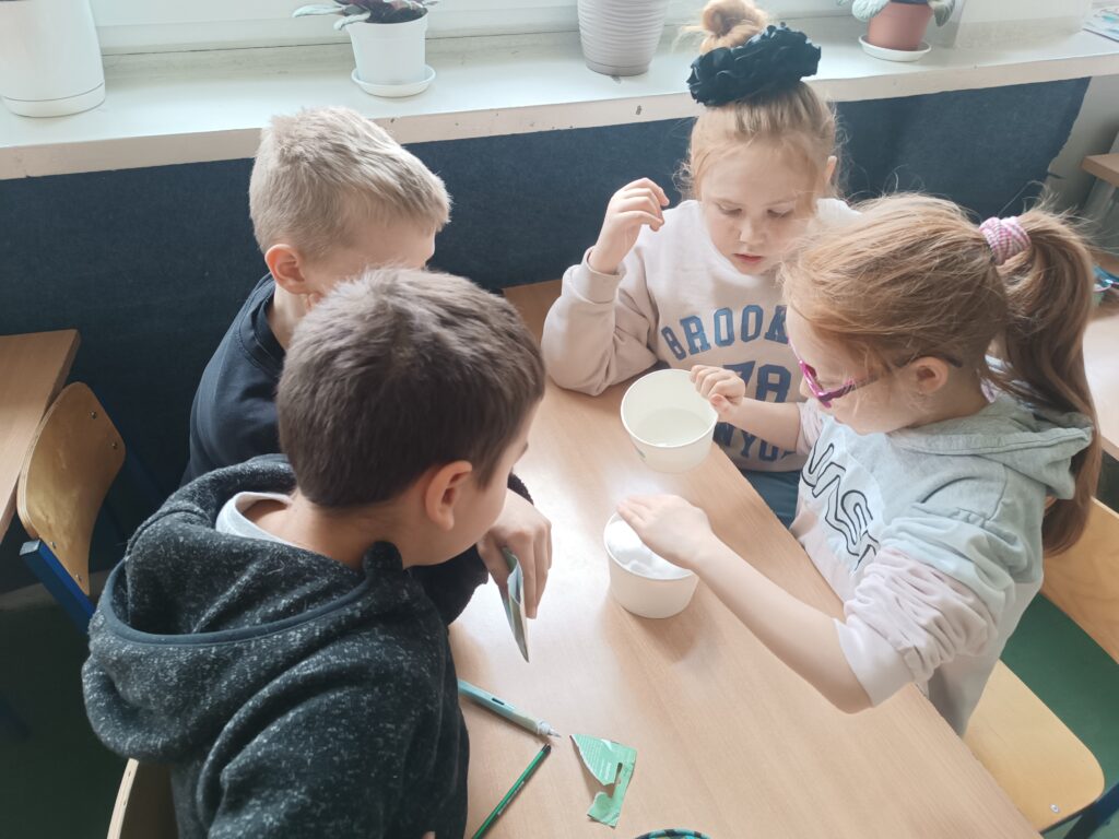 uczniowie siedzą grupą przy ławce, wsadzają ziarenka rzeżuchy do kubeczków  