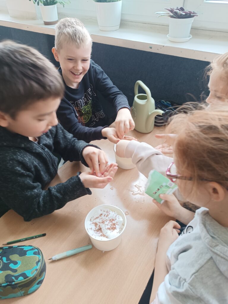 uczniowie siedzą grupą przy ławce, wsadzają ziarenka rzeżuchy do kubeczków  