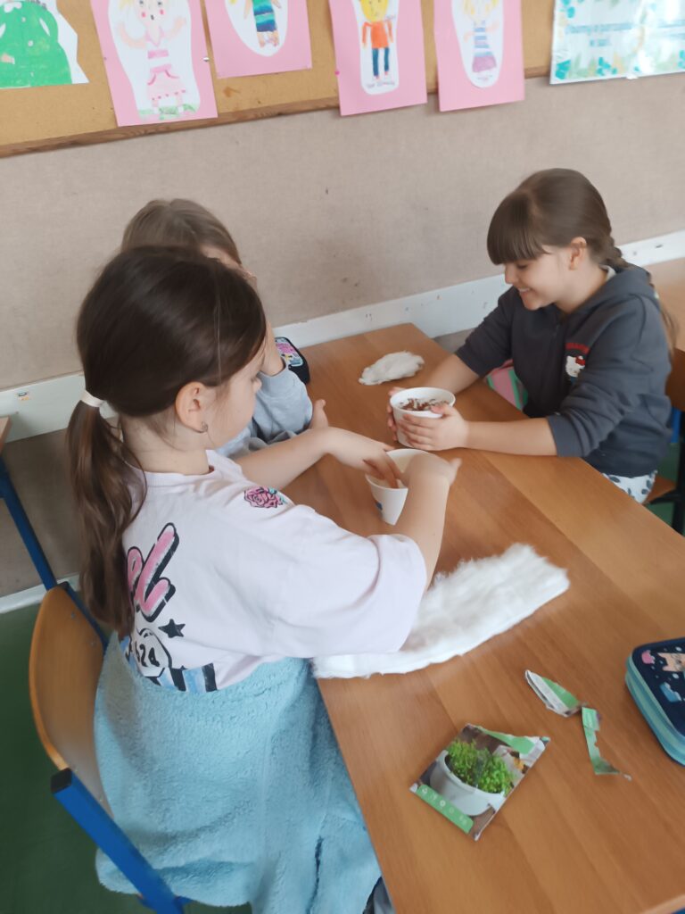 uczniowie siedzą grupą przy ławce, wsadzają ziarenka rzeżuchy do kubeczków  