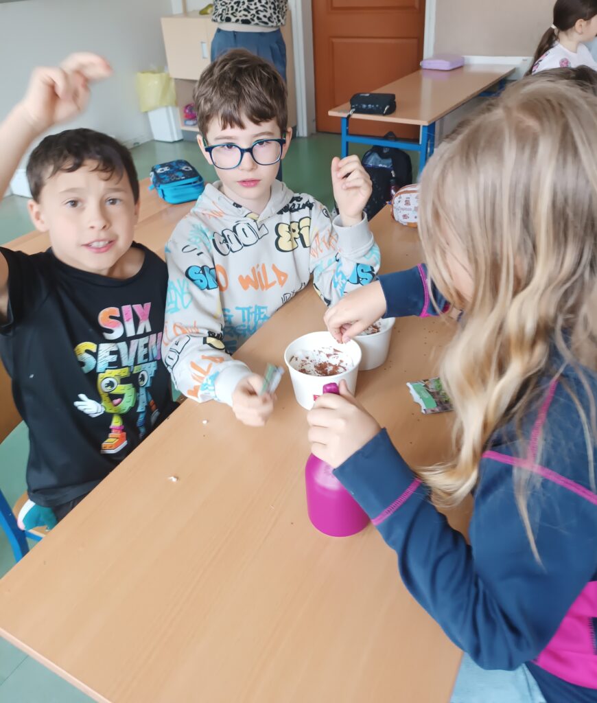 uczniowie siedzą grupą przy ławce, wsadzają ziarenka rzeżuchy do kubeczków  