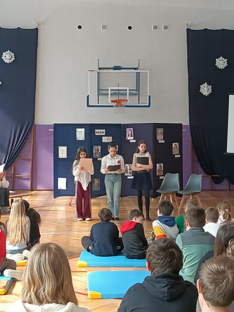 troje uczniów przemawia, na dole siedzą dzieci widać ich plecy