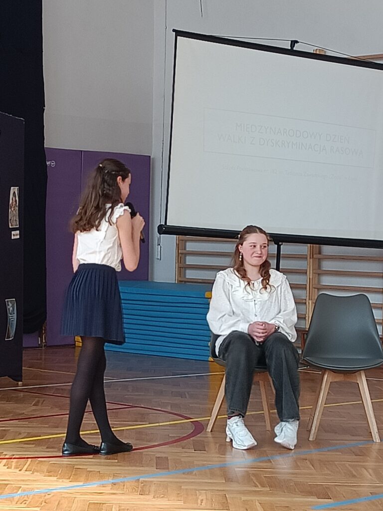 dwie dziewczynki odgrywają scenkę, jedna stoi i mówi, druga siedzi na krześle 