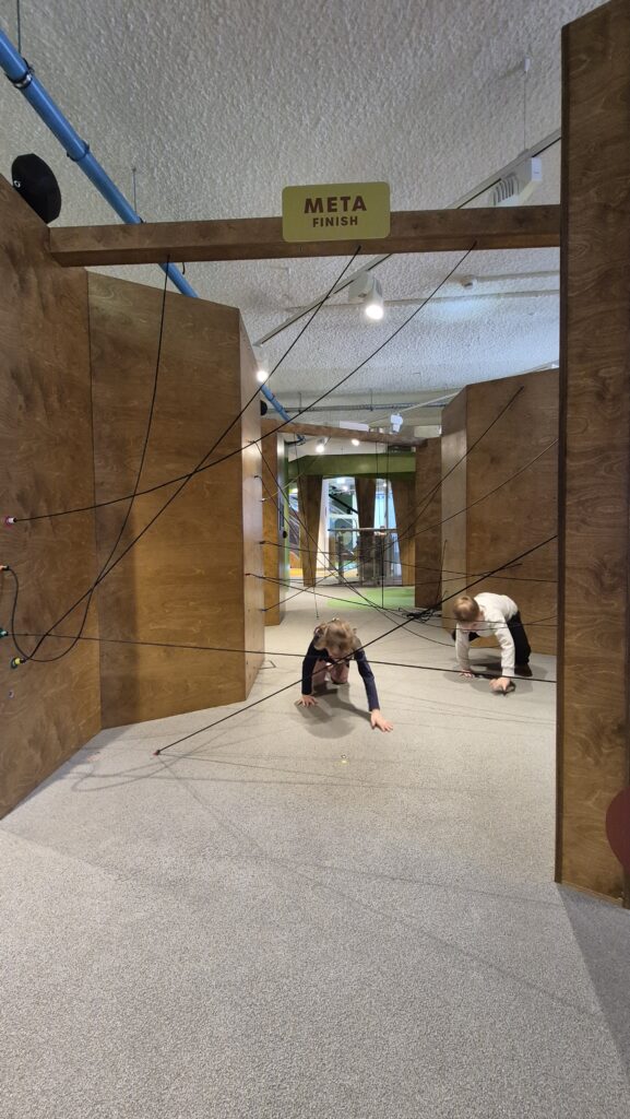 Uczniowie na czworaka pokonują tor przeszkód z czarnych linek przyczepionych do ścian.