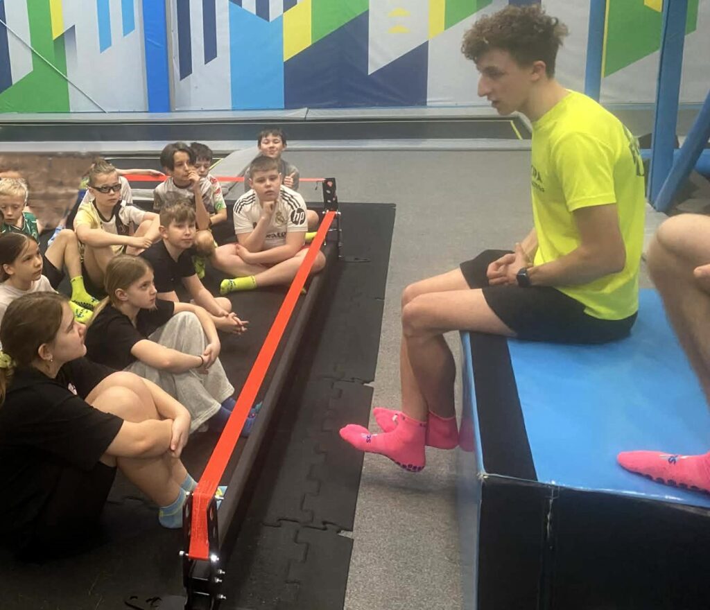 dzieci siedzą w sportowych strojach na podłodze i słuchają pana prowadzącego zajęcia w Saltosie