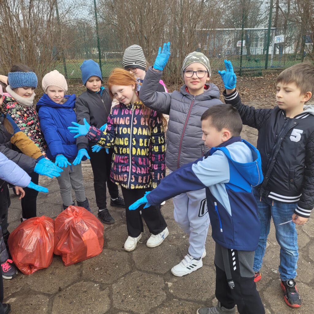 Na zdjęciu widać grupę uczniów w rękawiczkach ochronnych, zaangażowanych w sprzątanie terenu i pokazujących efekty swojej pracy.
