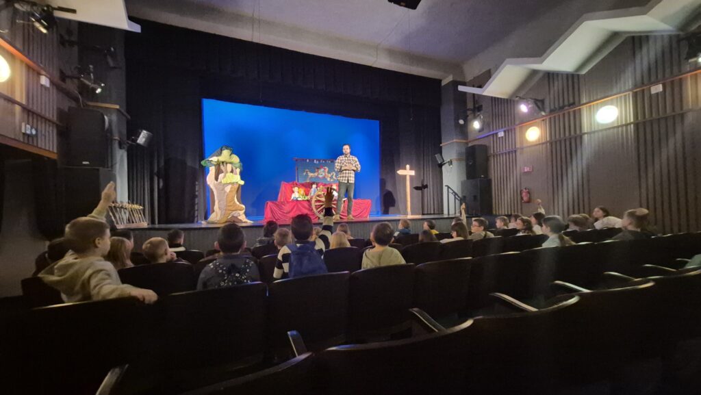 zdjęcie przedstawia dzieci od tyłu siedzące na widowni i na scenie aktora