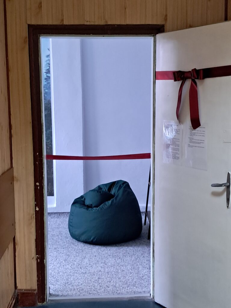 Wejście do Pokoju Wyciszeń. Zza wstęgi widoczny jest puf do wypoczynku w czasie przerw. Na drzwiach widoczna jest kokarda. W wejściu widać wstęgę do przecięcia podczas uroczystości.