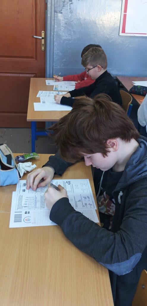 Uczniowie siedzą w klasie w ławkach nad arkuszami konkursowymi.