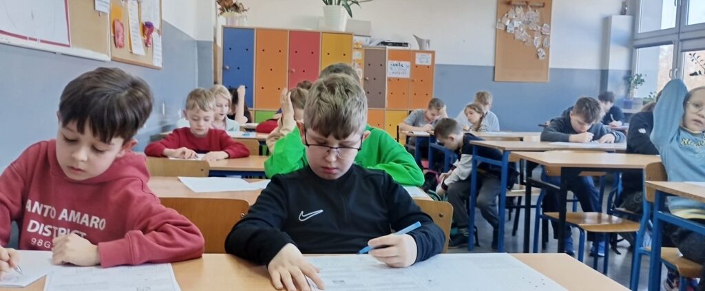 Uczniowie siedzą w klasie w ławkach nad arkuszami konkursowymi.