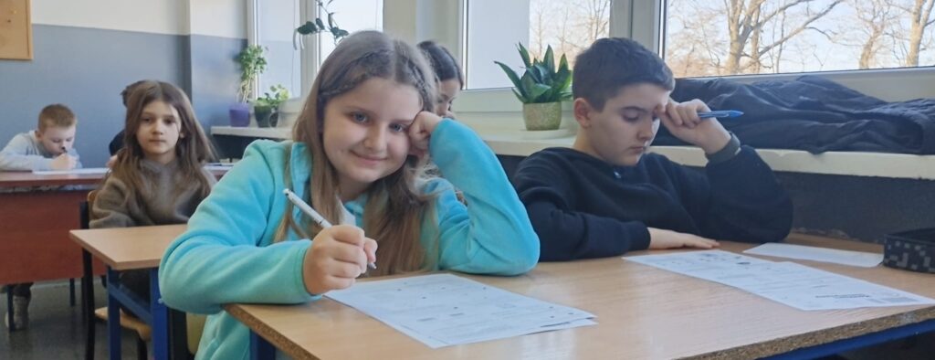 Uczniowie siedzą w klasie w ławkach nad arkuszami konkursowymi.