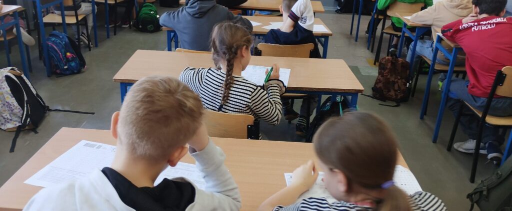 Uczniowie siedzą w klasie w ławkach nad arkuszami konkursowymi.