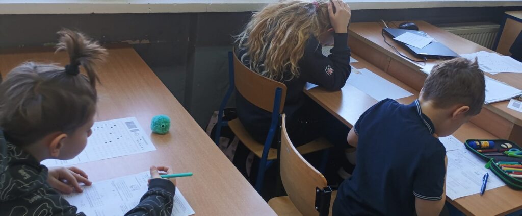 Uczniowie siedzą w klasie w ławkach nad arkuszami konkursowymi.