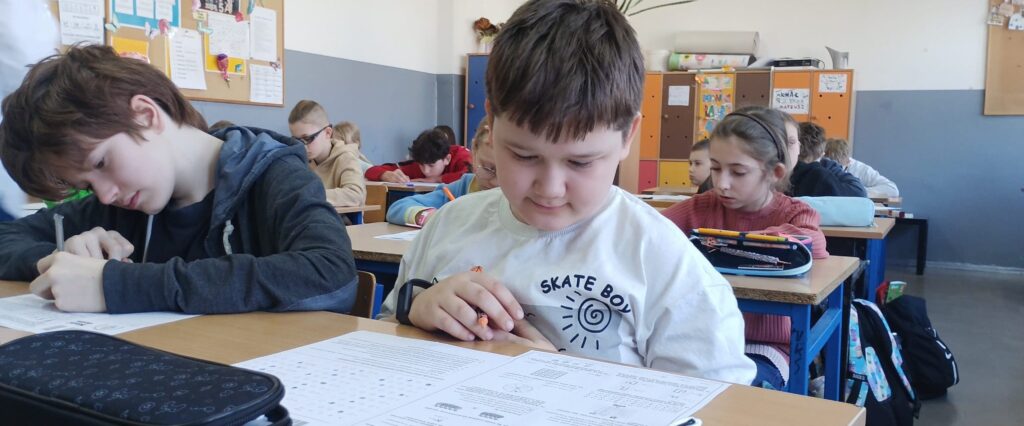 Uczniowie siedzą w klasie w ławkach nad arkuszami konkursowymi.