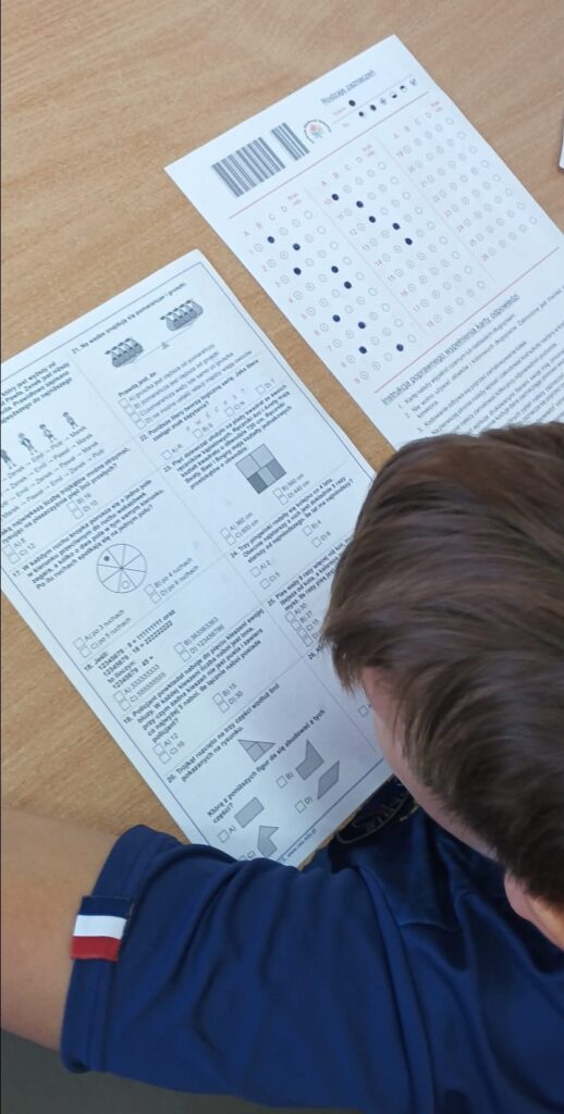 Uczniowie siedzą w klasie w ławkach nad arkuszami konkursowymi.