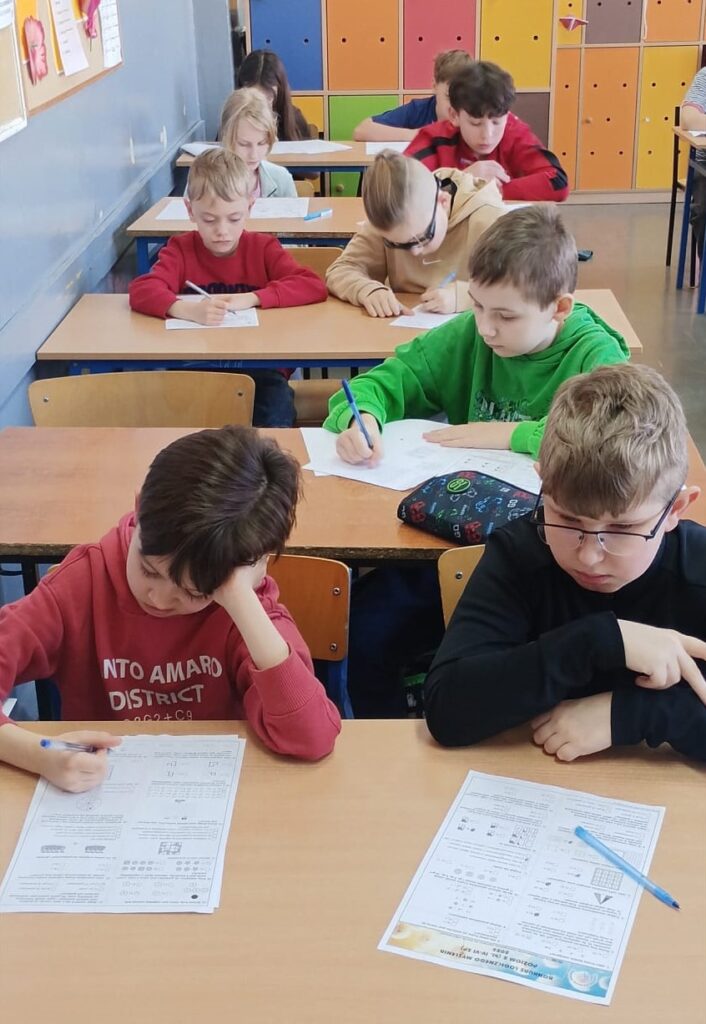 Uczniowie siedzą w klasie w ławkach nad arkuszami konkursowymi.