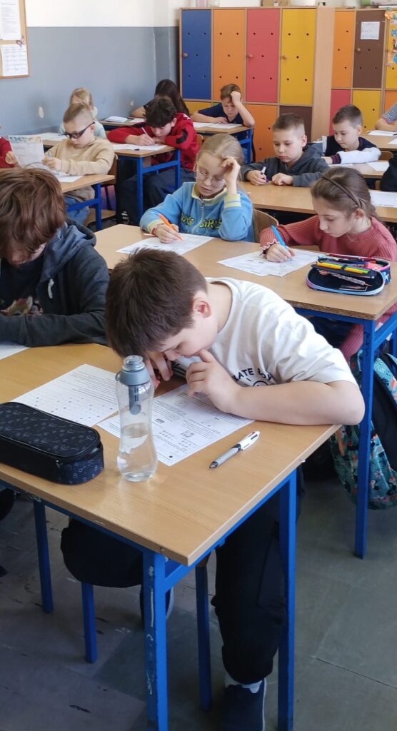 Uczniowie siedzą w klasie w ławkach nad arkuszami konkursowymi.