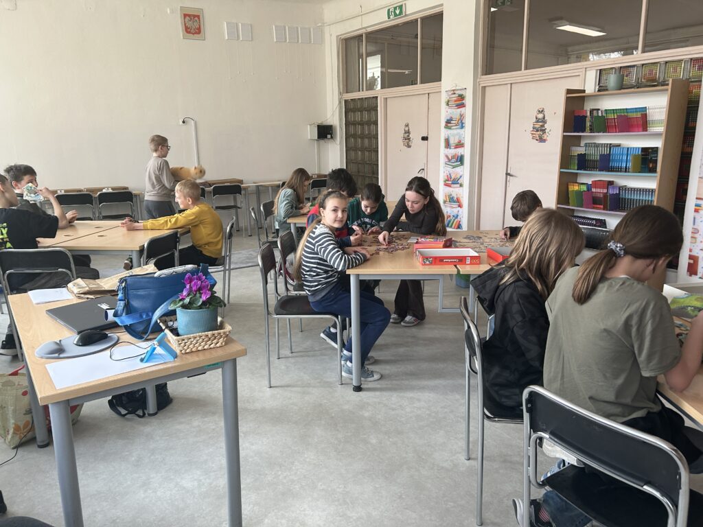 widok na bibliotekę. Przy stolikach siedzą dzieci i grają w gry