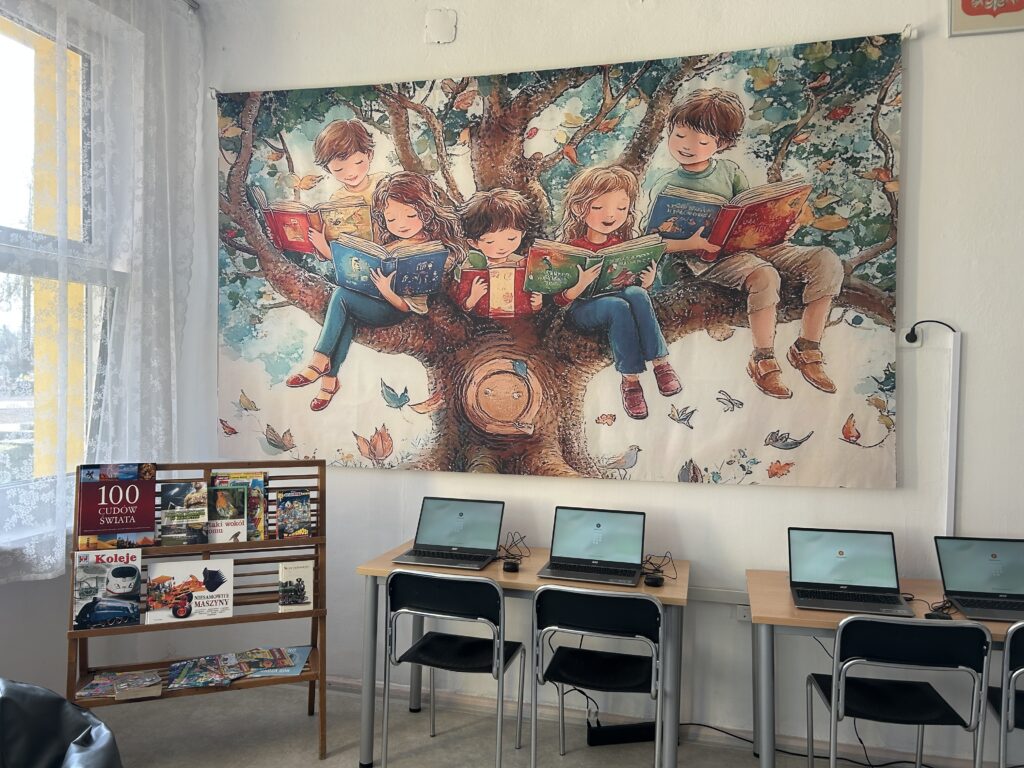 widok na ścianę z dużym, kolorowym banerem (drzewo, gdzie siedzą dzieci i czytają książki). Na stolikach laptopy.