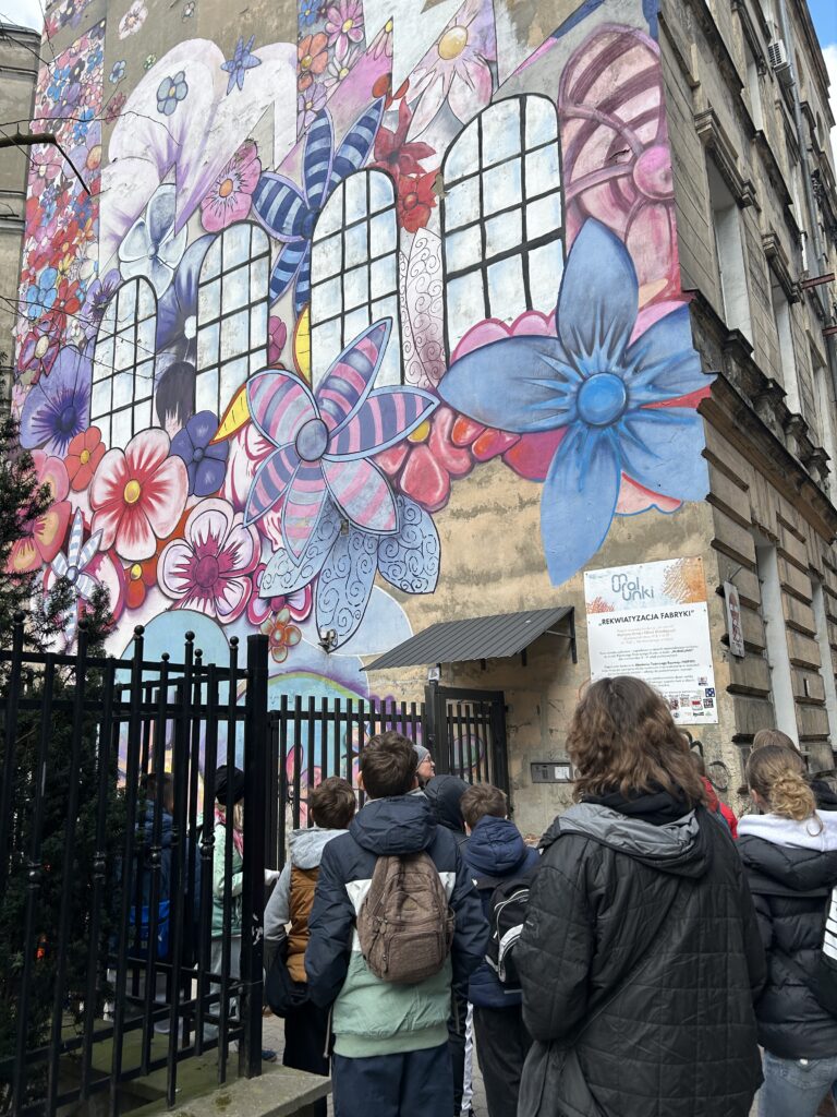 kamienica przy ul. Próchnika. Na ścianie namalowane kolorowe kwiaty. Uczniowie stoją przodem do kamienicy i przyglądają się muralowi