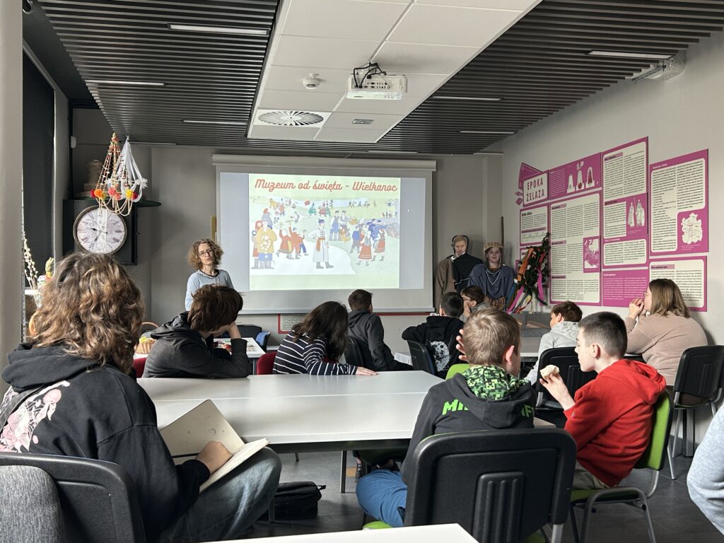uczniowie siedzą przy stolikach w sali edukacyjnej  w Muzeum Archeologicznym i Etnograficznym i oglądają prezentację zatytułowaną Muzeum od świeta Wielkanoc. Przodem do uczniów stoi wygłaszająca prelekcję przewodniczka z muzeum
