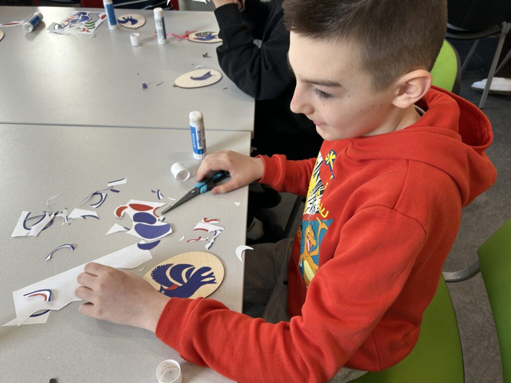 chłopiec siedzi przy stoliku. Przed nim  leży jego praca - drewniany kształt jajka, na nim przyklejony kolorowy kogucik