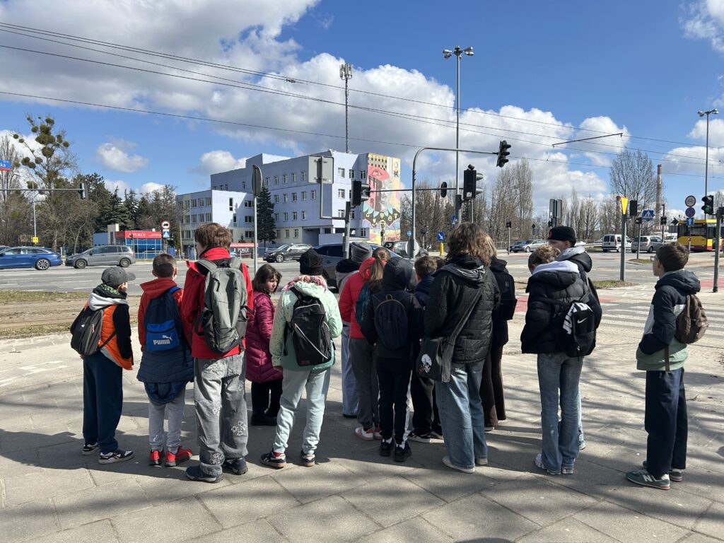 uczniowie stoją tyłem do fotografa, przodem do ulicy Aleksandrowskiej i z daleka podziwiają mural na Spółdzielni "Pokój"