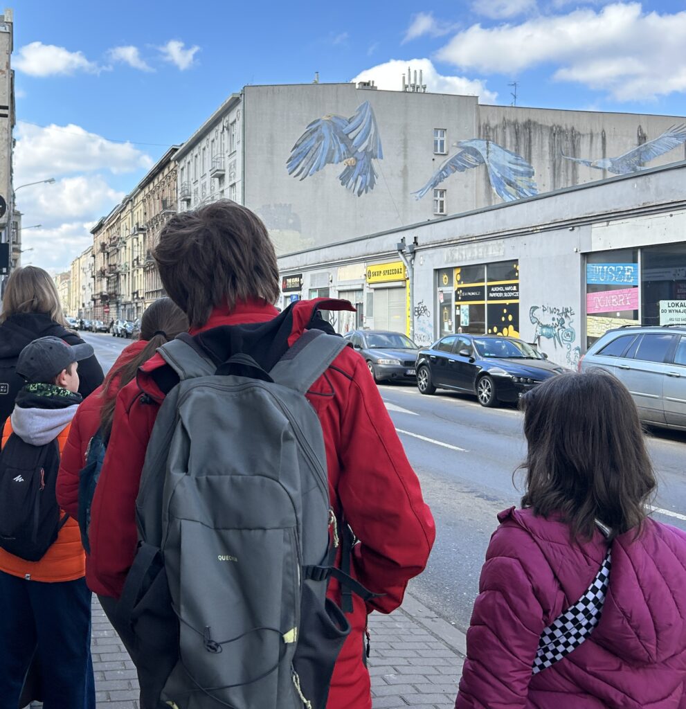 uczniowie stoją tyłem do fotografa, przodem do ulicy Próchnika i podziwiają mural na kamienicy - trzy błękitne orły