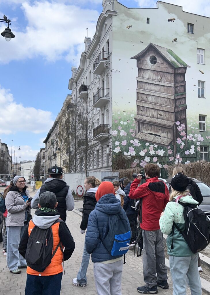 uczniowie uczniowie stoją tyłem do fotografa, przodem do al. 1 Maja  i podziwiają mural na kamienicy - "Ul z pszczołami"