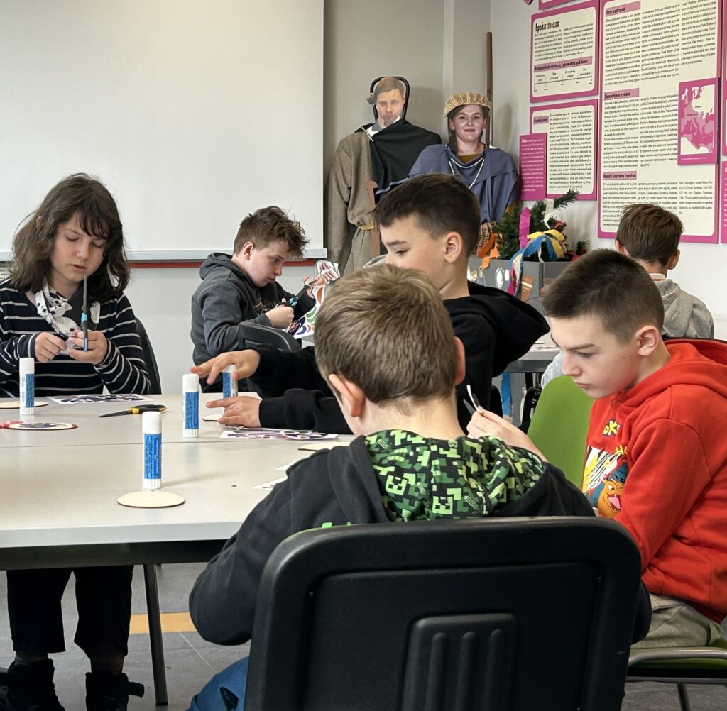 uczniowie siedzą przy stolikach w sali edukacyjnej  w Muzeum Archeologicznym i Etnograficznym i wycinają elementy koguta na ozdobę świąteczną