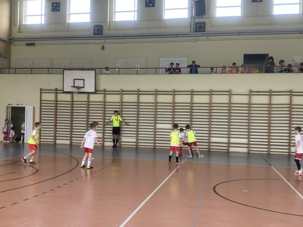 kolejne uczniowie grają w piłkę nożną na hali, drużyny w żółtych i białych koszulkach