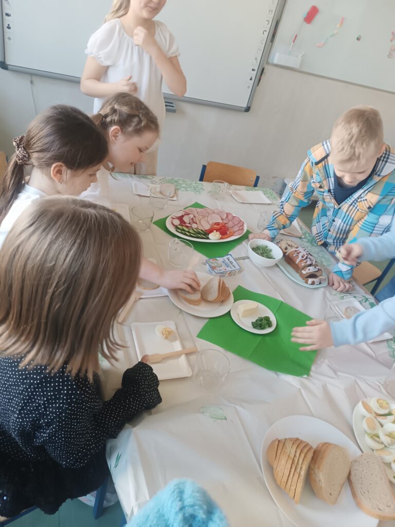 dzieci siedzą przy ławeczkach przystrojonych obrusem białym, wędliną, warzywami, rzeżuchą , ciastem i jajkami.