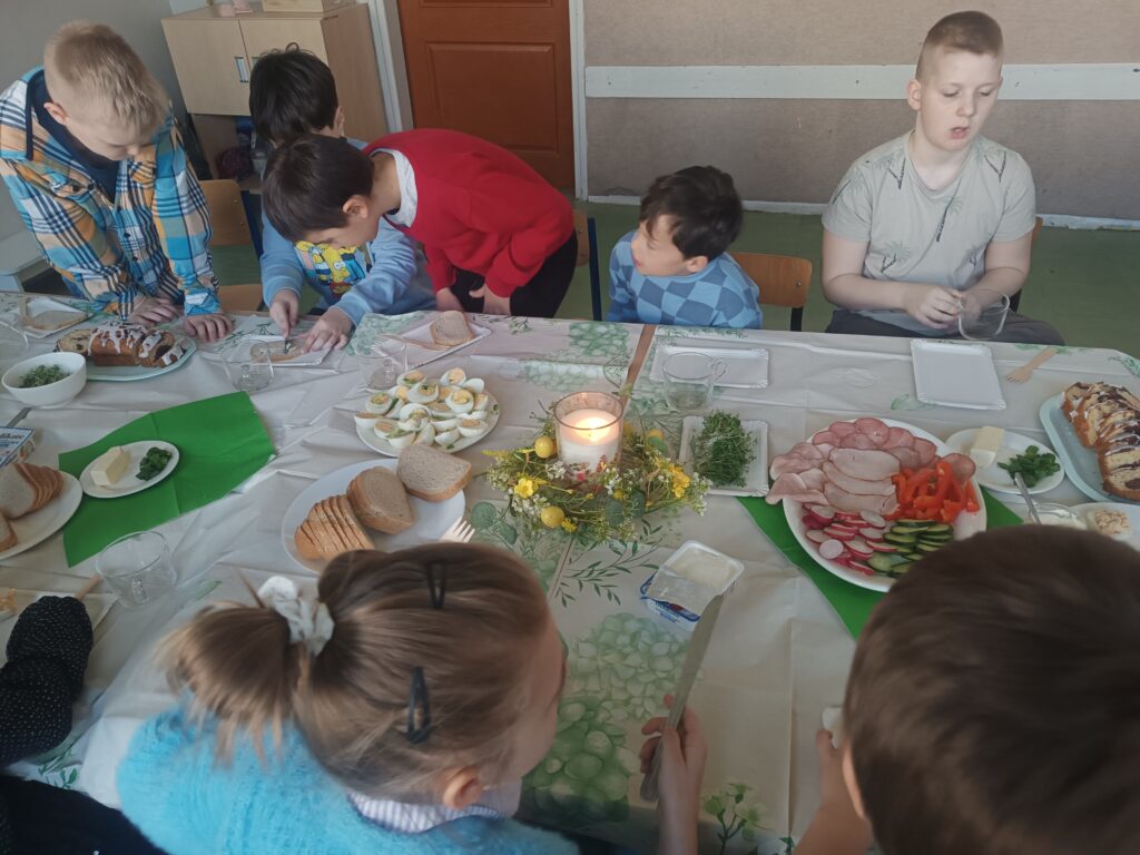 dzieci siedzą przy ławeczkach przystrojonych obrusem białym, wędliną, warzywami, rzeżuchą , ciastem i jajkami.