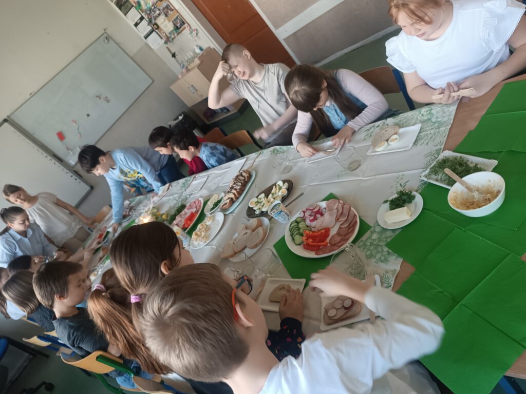 dzieci siedzą przy ławeczkach przystrojonych obrusem białym, wędliną, warzywami, rzeżuchą , ciastem i jajkami.