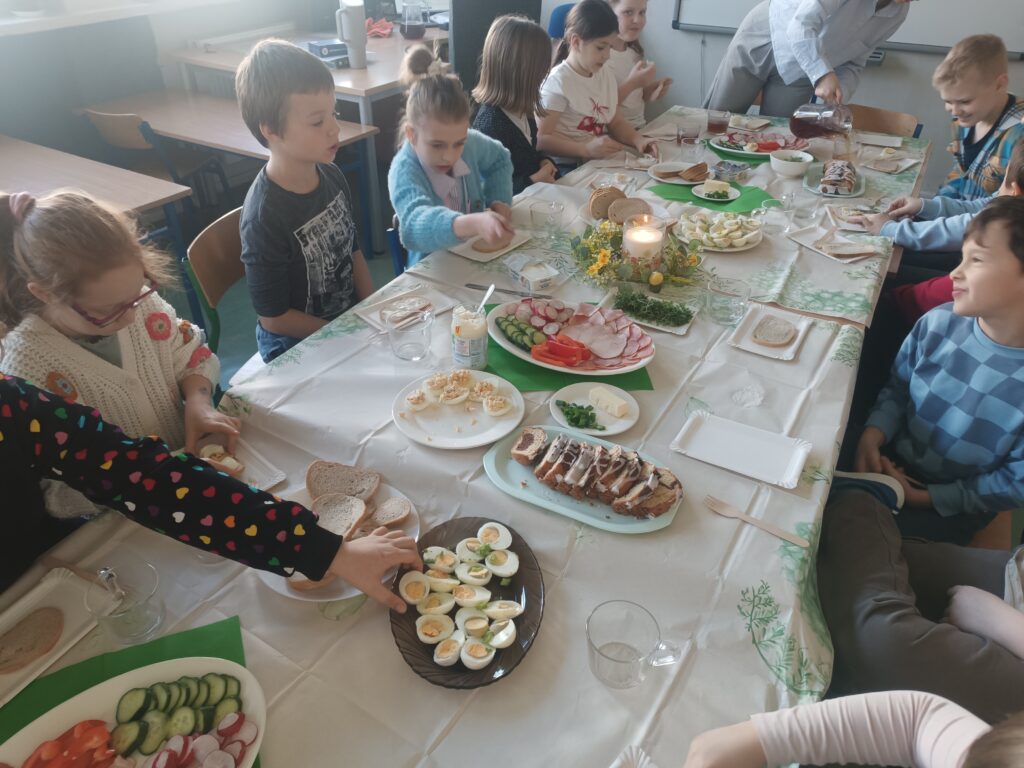 dzieci siedzą przy ławeczkach przystrojonych obrusem białym, wędliną, warzywami, rzeżuchą , ciastem i jajkami.