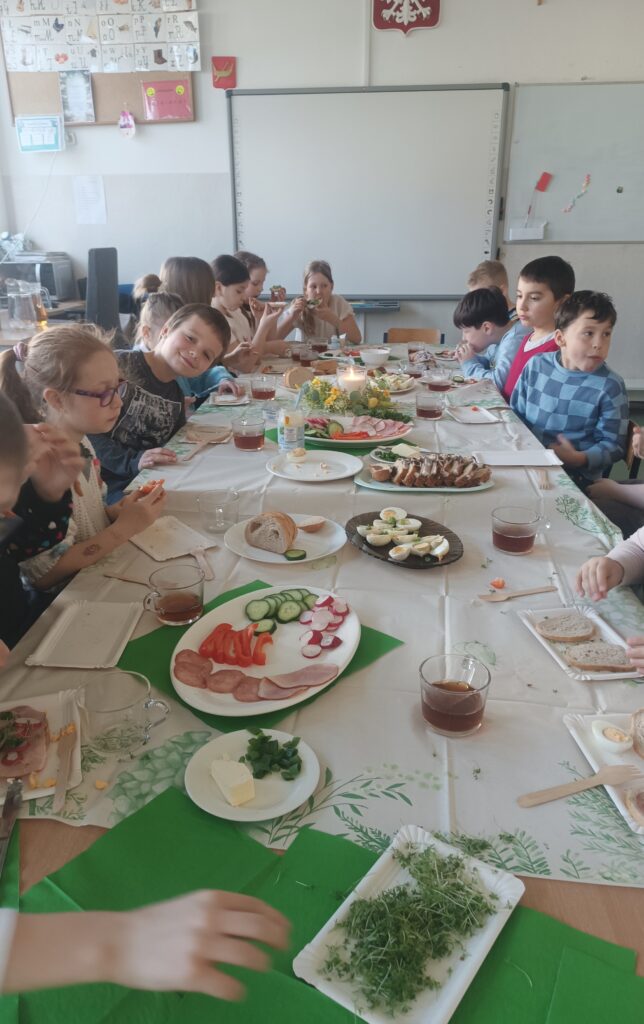 dzieci siedzą przy ławeczkach przystrojonych obrusem białym, wędliną, warzywami, rzeżuchą , ciastem i jajkami.