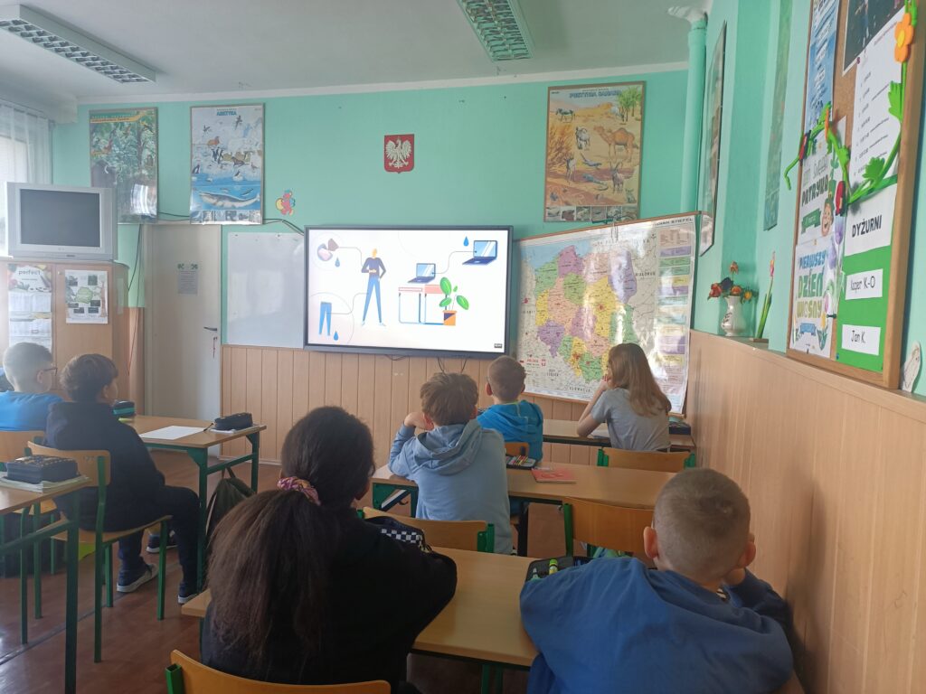 dzieci siedzą w ławkach, widzimy ich od tyłu, oglądają film wyświetlony na ekranie interaktywnym 