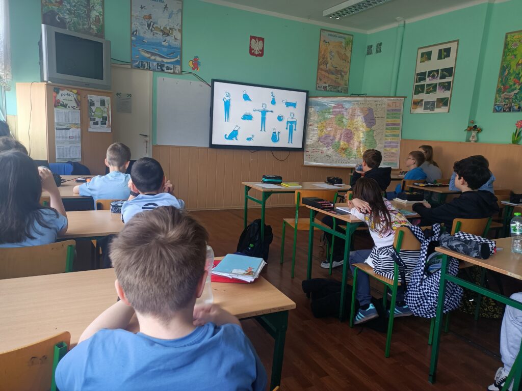 dzieci siedzą w ławkach, widzimy ich od tyłu, oglądają film wyświetlony na ekranie interaktywnym 