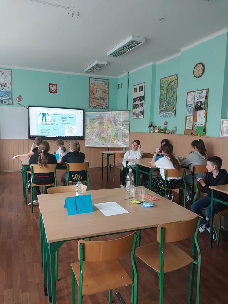 dzieci siedzą w ławkach, widzimy ich od tyłu, oglądają film wyświetlony na ekranie interaktywnym 