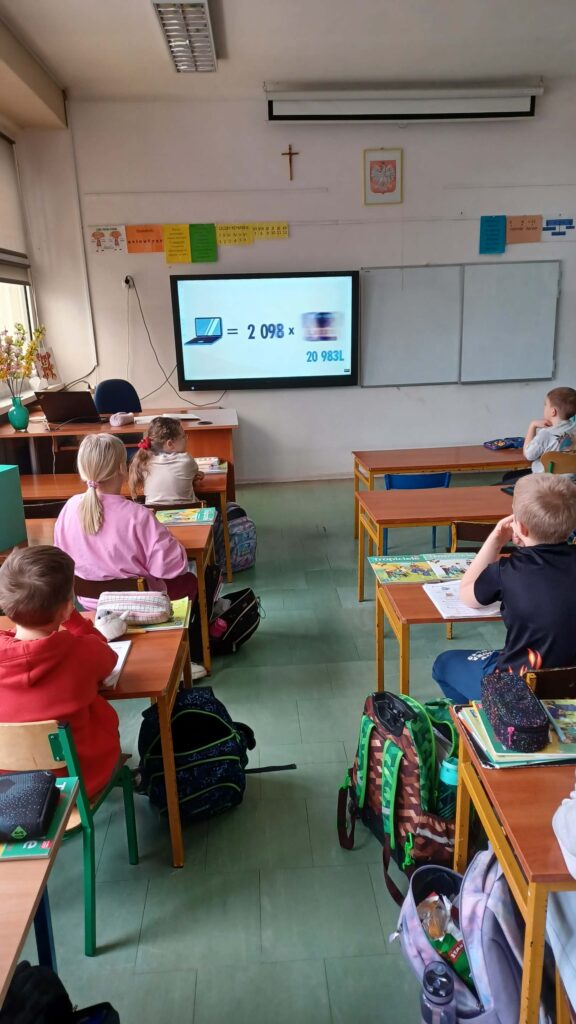 dzieci siedzą w ławkach, widzimy ich od tyłu, oglądają film wyświetlony na ekranie interaktywnym 